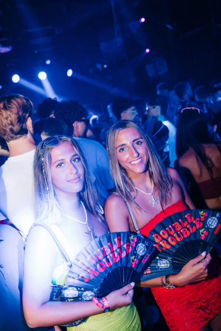Two girls holding Double Trouble fans and smiling on the packed dancefloor at Toy Room Super Club Night in Malta