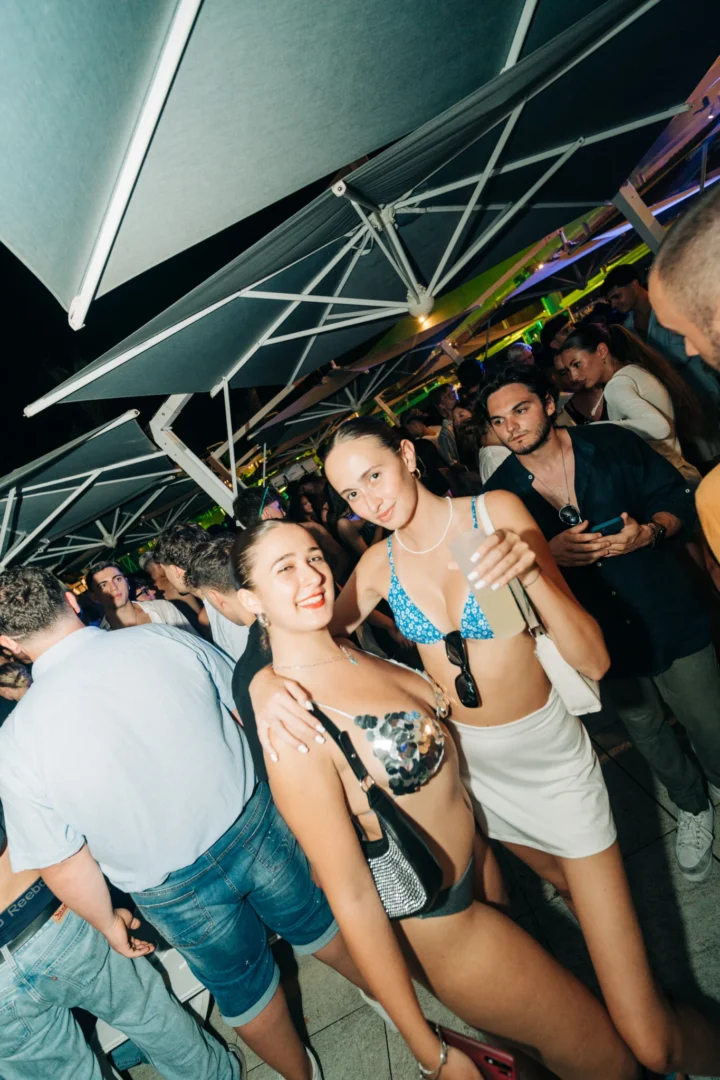 Girls smiling with cocktails under the umbrellas at Cafe Del Mar Malta, packed dance floor, late night tunes and neon lights
