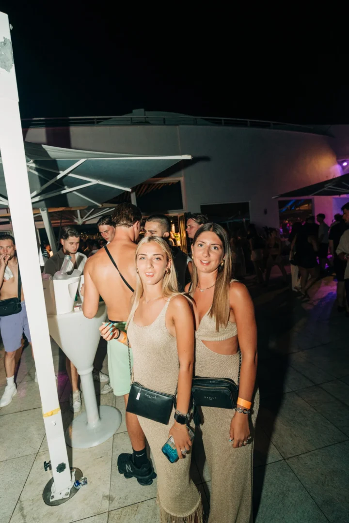 Two girls posing by the bar at Cafe Del Mar Malta night party, glowing lights, cocktails flowing and crowds packing the terrace