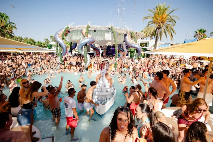 Performer standing on a drinks boat in the pool at O Beach Ibiza with a packed crowd, yellow parasols and giant tentacles above the stage