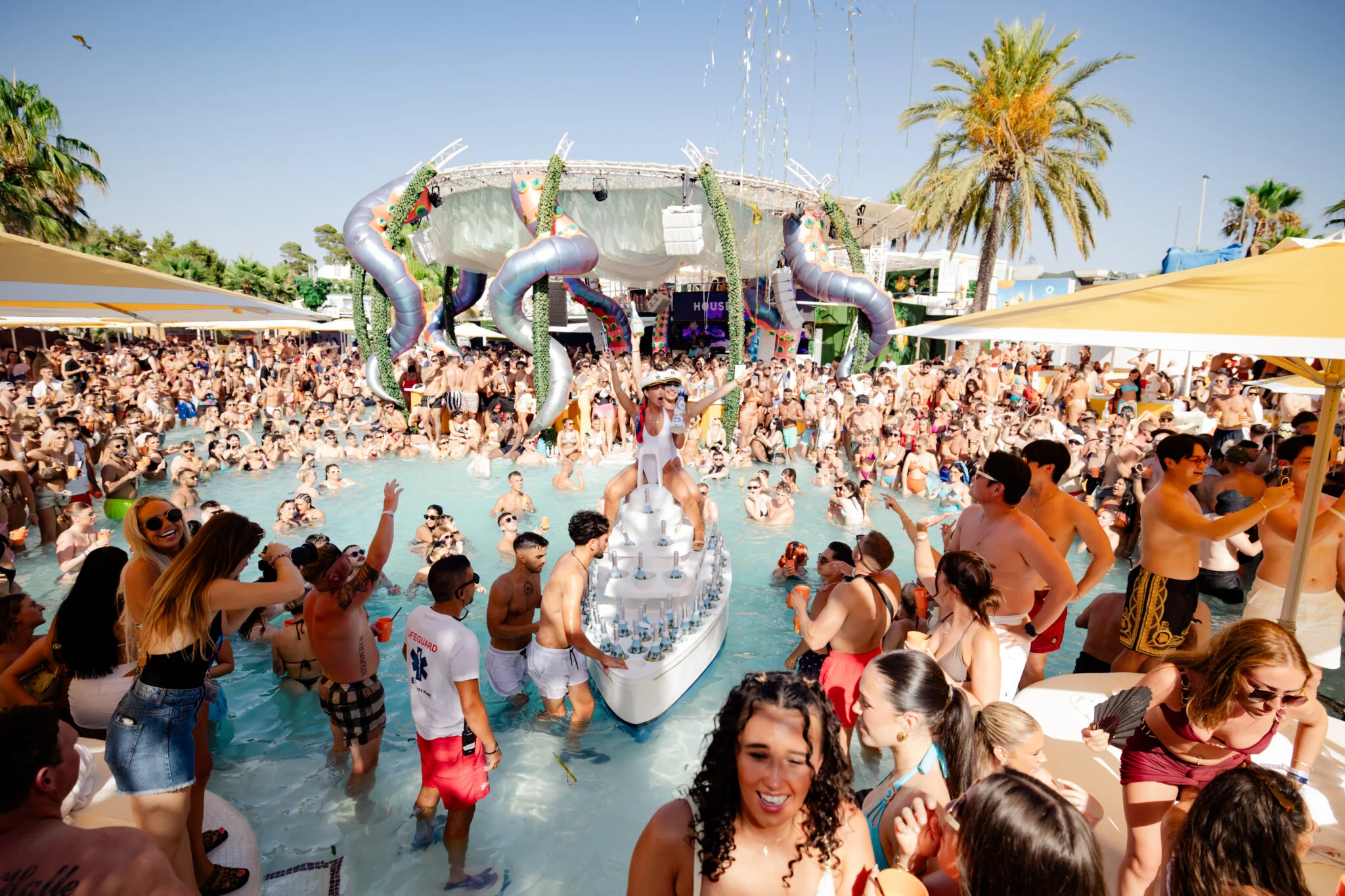 Performer standing on a drinks boat in the pool at O Beach Ibiza with a packed crowd, yellow parasols and giant tentacles above the stage