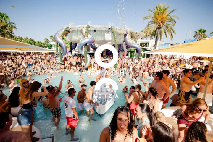 Packed crowd in and around the pool at O Beach Ibiza with palm trees, stage decor and a drinks boat in the water