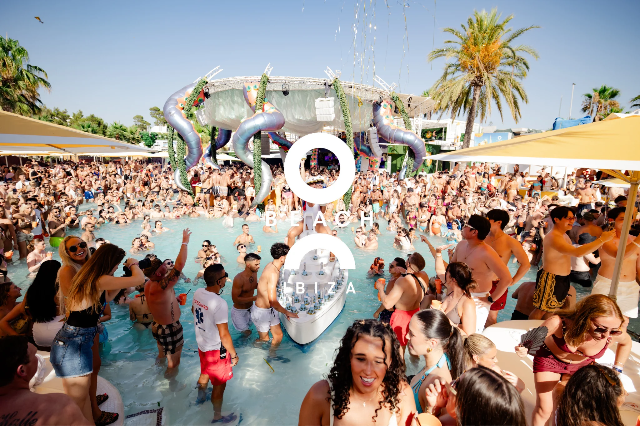 Packed crowd in and around the pool at O Beach Ibiza with palm trees, stage decor and a drinks boat in the water