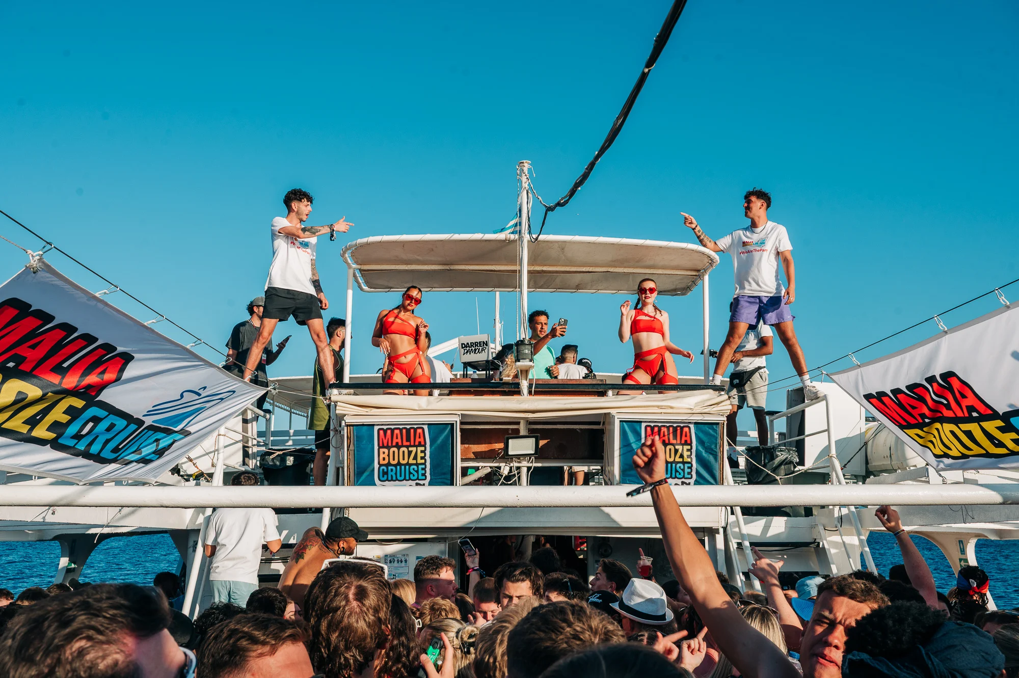 Performers in red dance on deck while DJs hype the crowd below – Malia Booze Cruise hits peak party mode under clear blue skies.