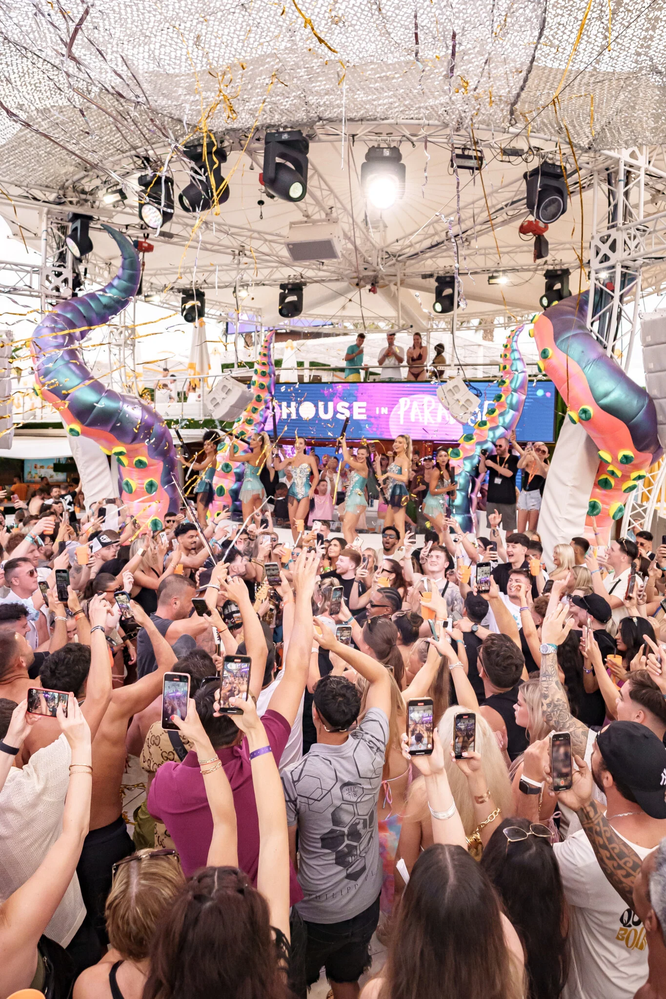 Packed crowd holding phones up in front of the stage at House in Paradise at O Beach Ibiza with dancers, confetti and giant octopus tentacles