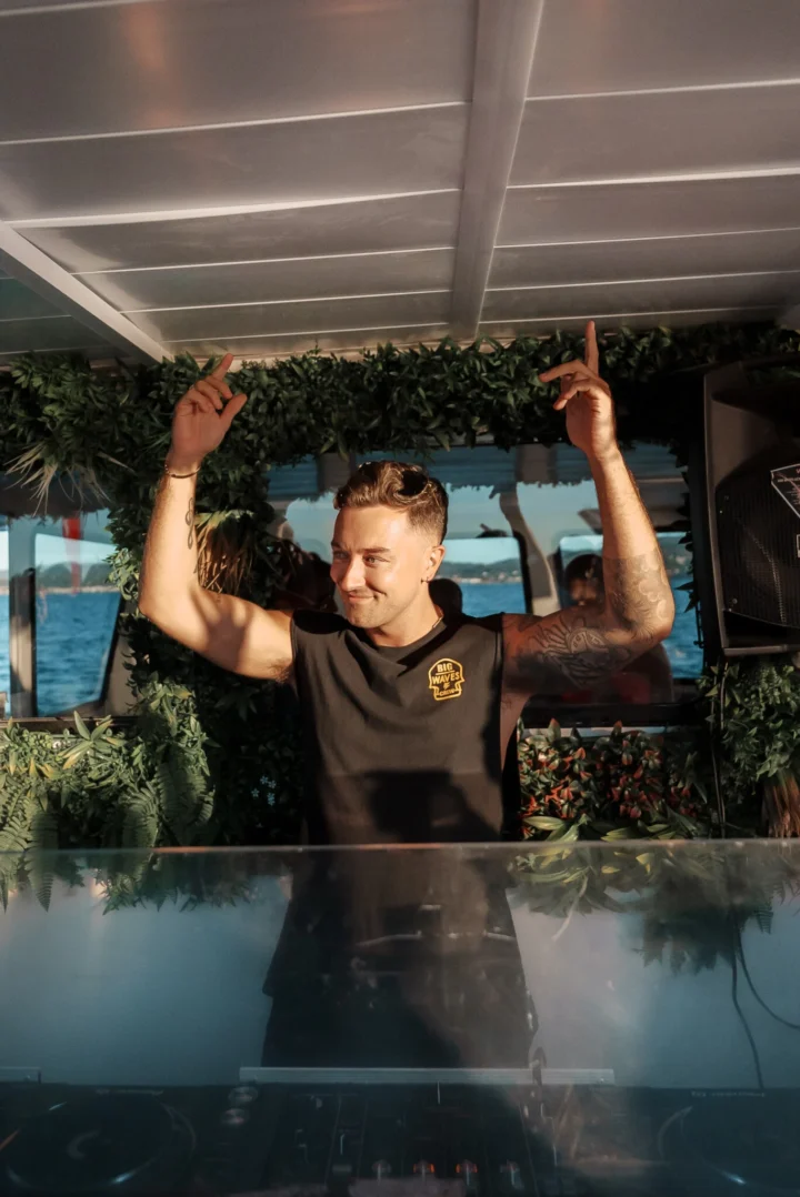 DJ with both hands raised behind the booth on a Pukka Up boat party in Ibiza with the sea visible through the windows