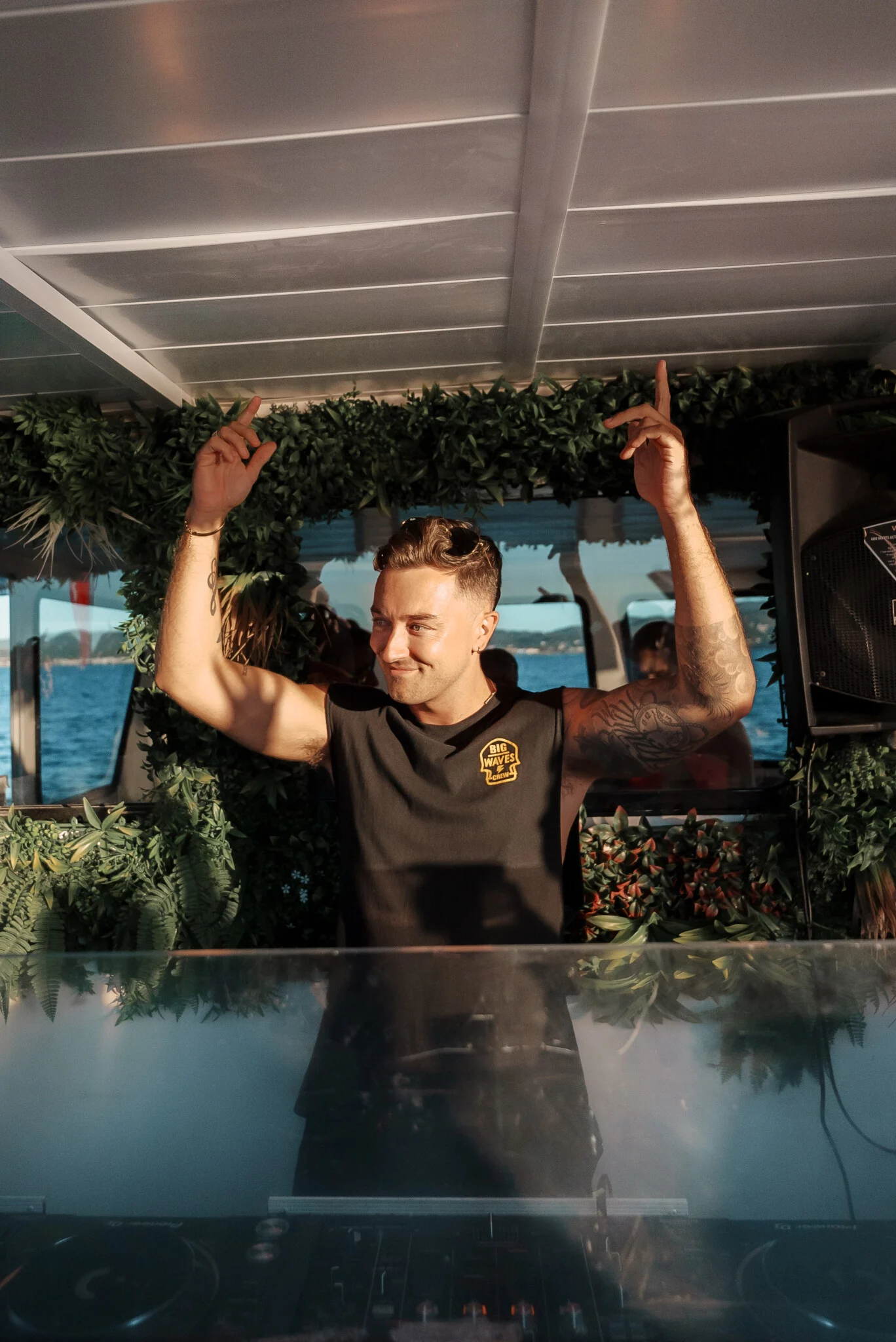 DJ with both hands raised behind the booth on a Pukka Up boat party in Ibiza with the sea visible through the windows