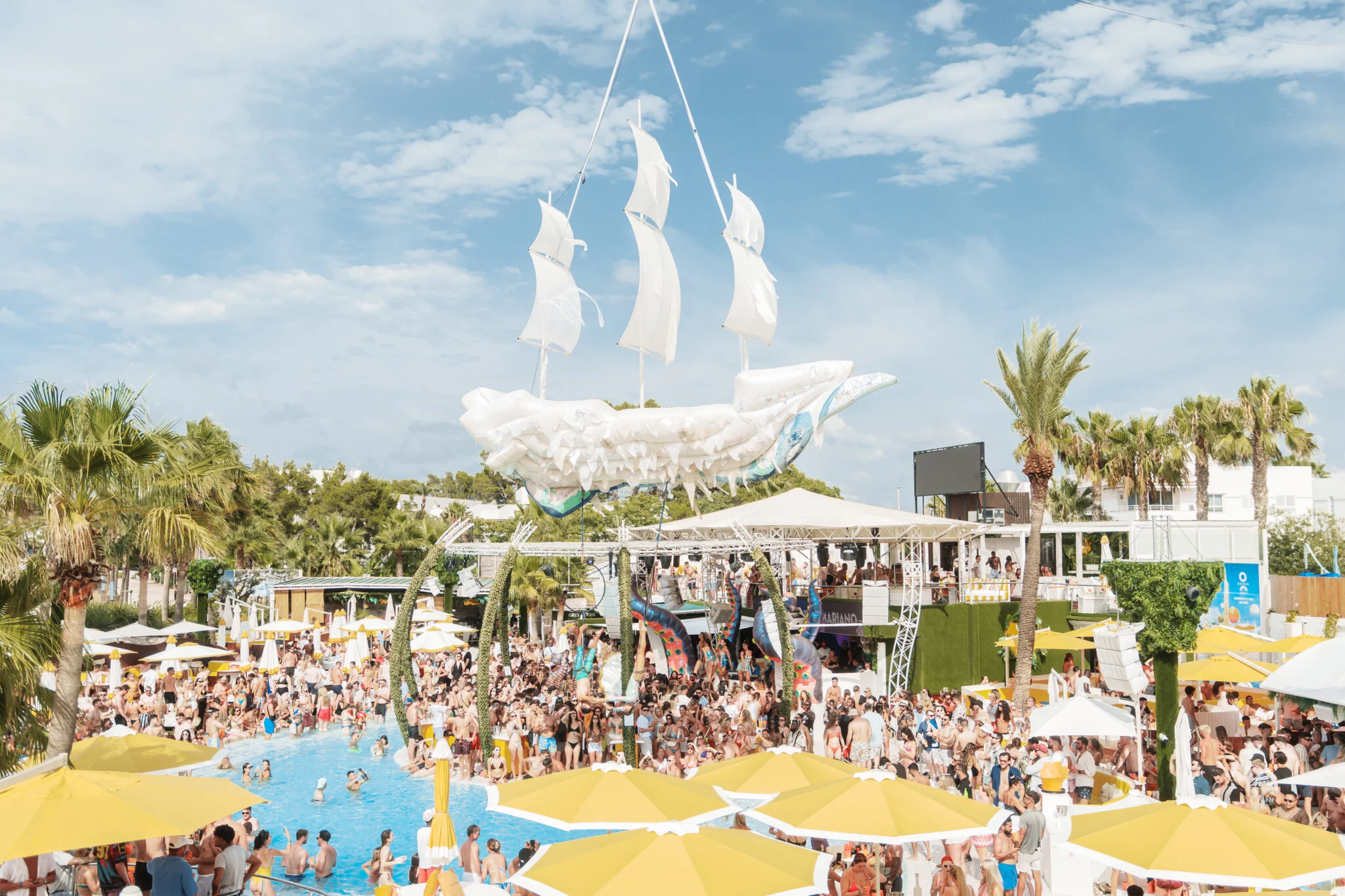 Wide view of O Beach Ibiza with the pool, packed crowd, yellow parasols and a large white ship decoration suspended above the venue