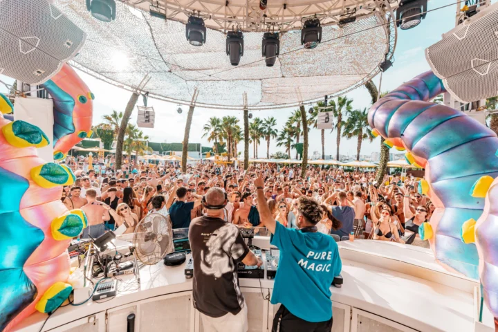 DJ booth view of a packed crowd with hands in the air at O Beach Ibiza framed by giant colourful tentacles and palm trees