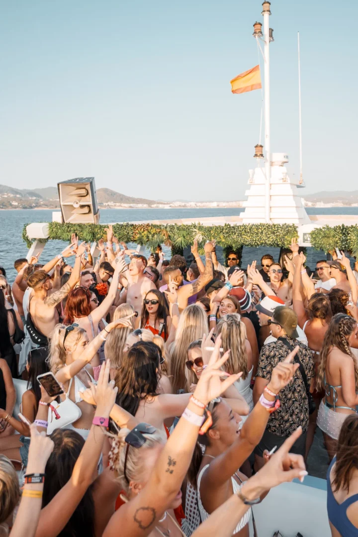 Packed crowd with hands in the air dancing on a Pukka Up boat party in Ibiza with the sea and coastline behind them