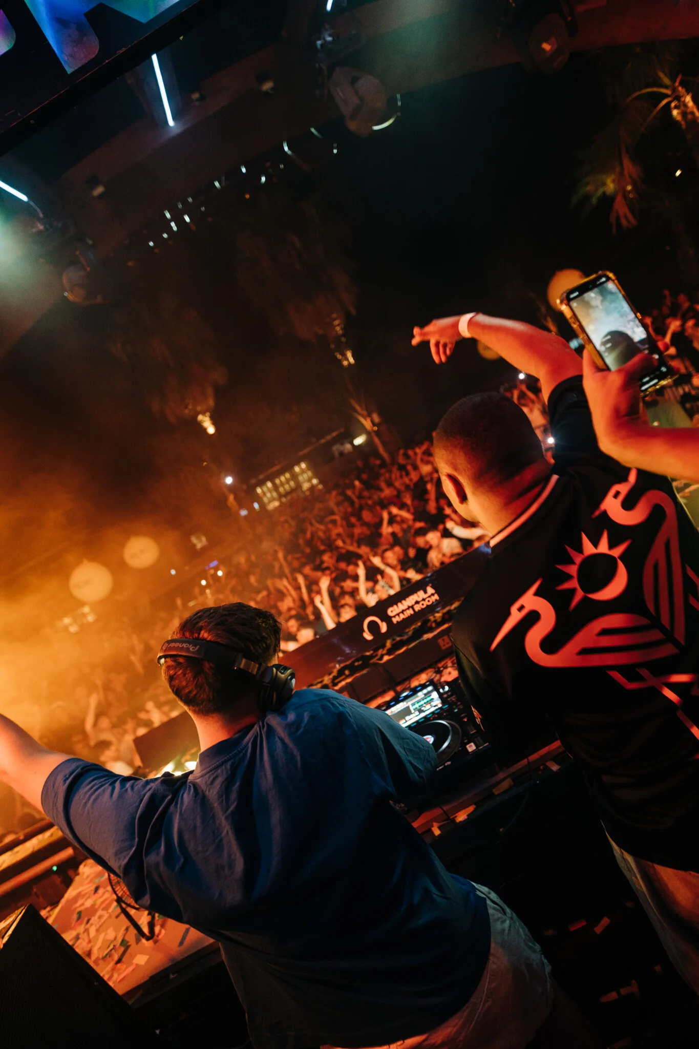 DJs performing at Gianpula Main Room Club Night in Malta, with one filming the crowd as orange smoke and stage lights fill the venue.