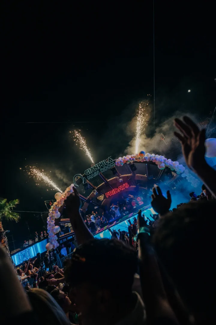 Raised hands in the crowd at Gianpula Main Room Club Night in Malta as fireworks burst above the stage and blue lights fill the venue.