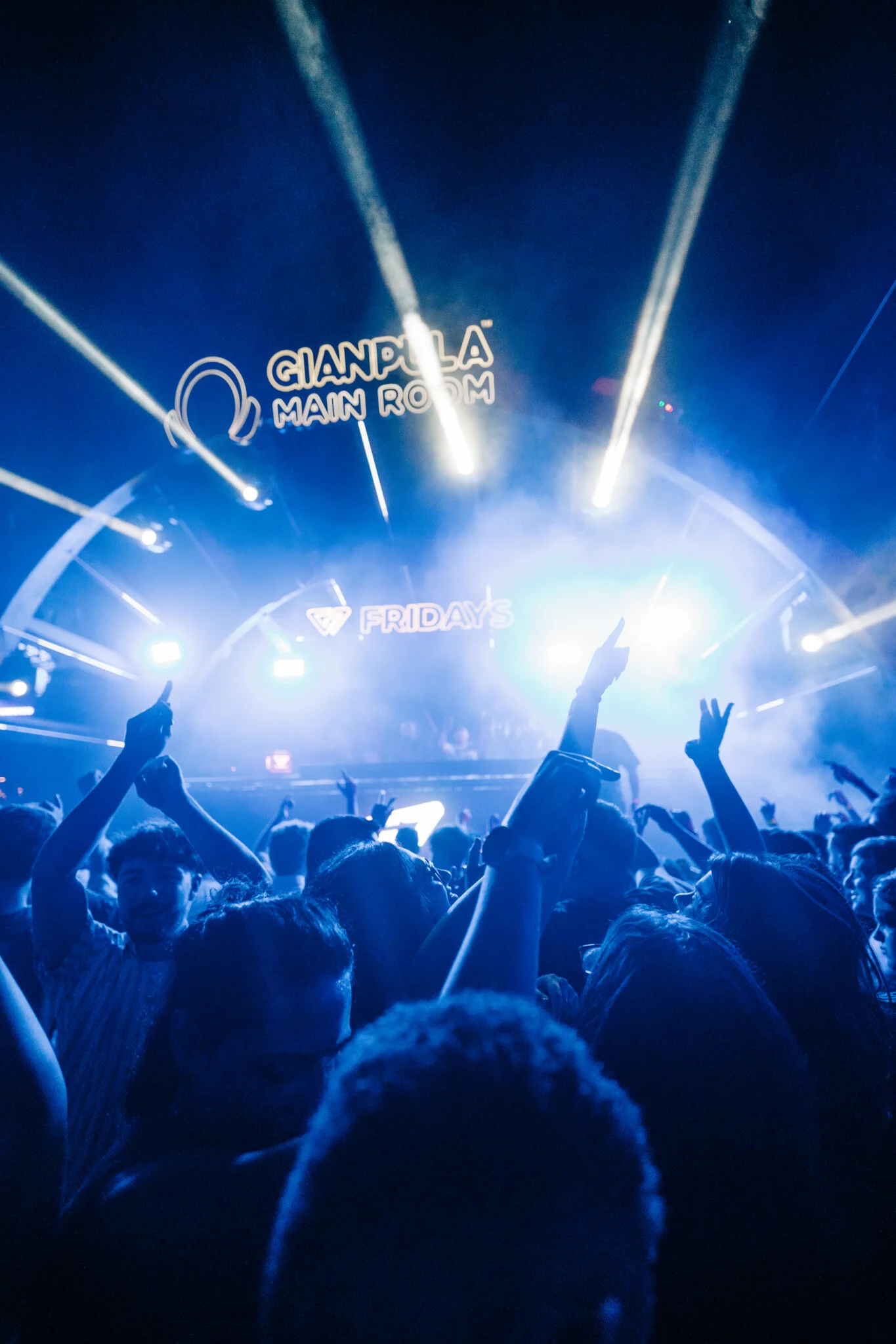 Crowd dancing at Gianpula Main Room Club Night in Malta beneath bright blue stage lights and smoke, with hands raised toward the DJ booth.