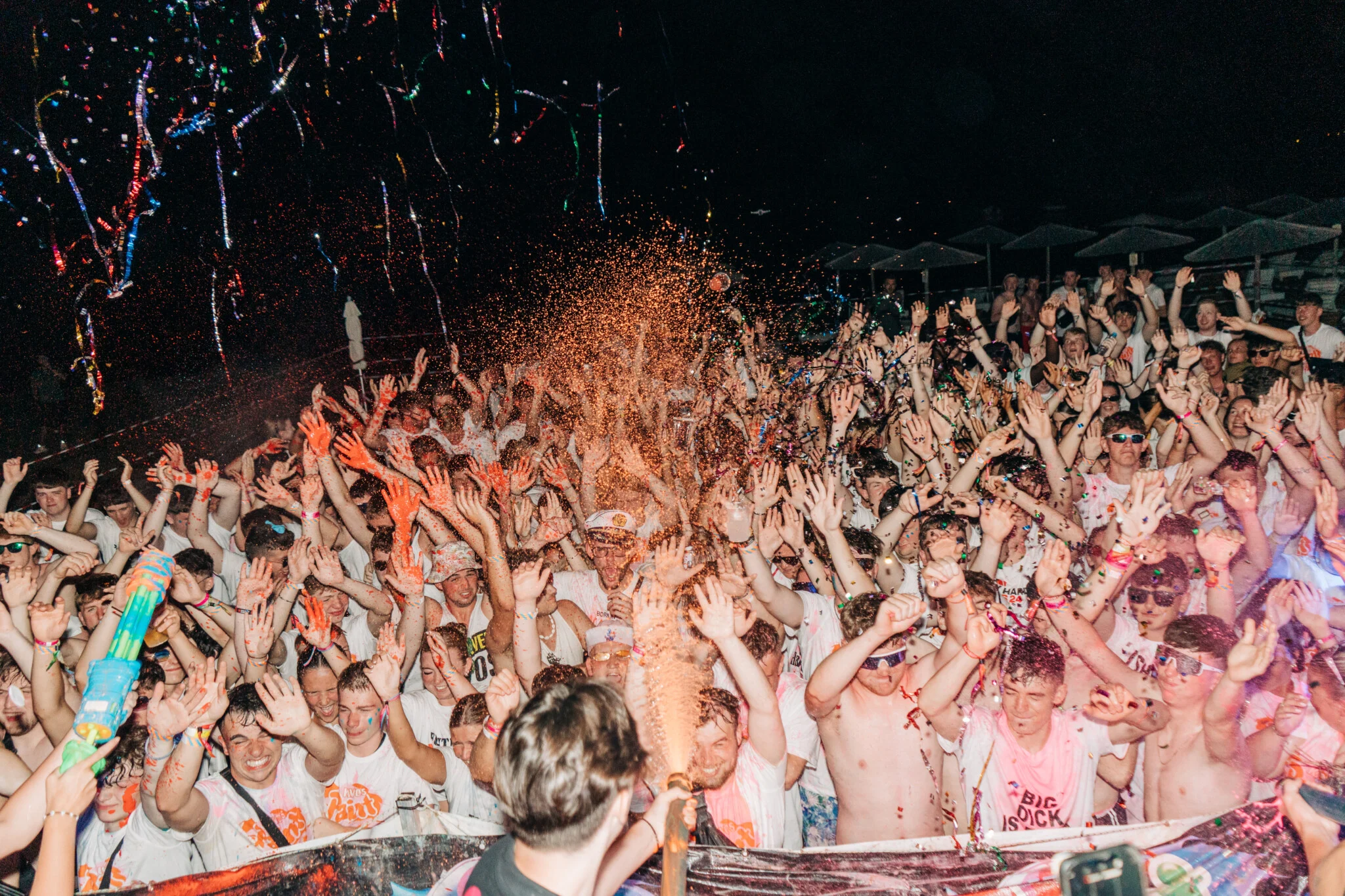 Massive crowd going wild at a Kavos paint party, hands in the air as paint sprays, music pumping and chaos erupting at night