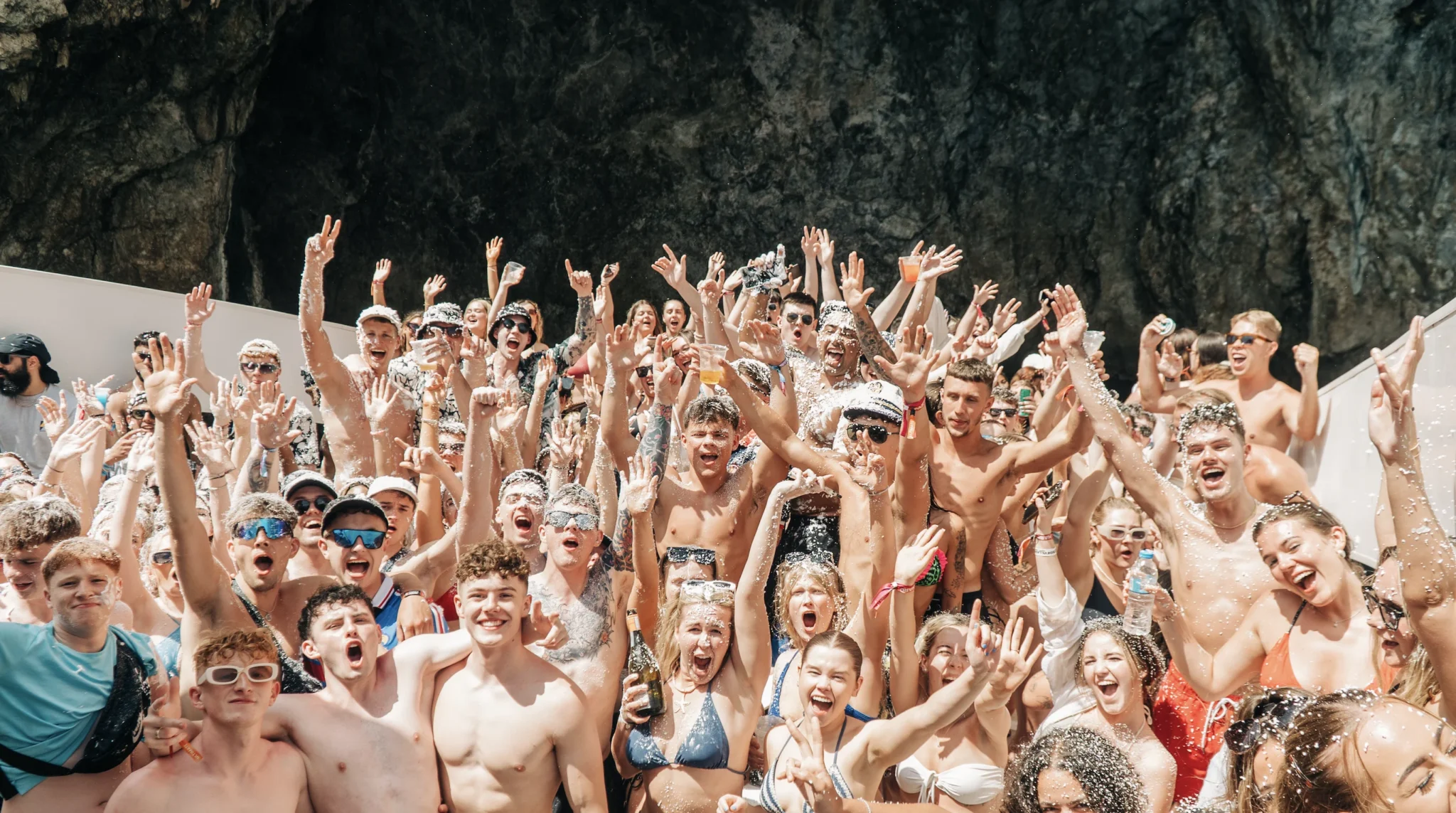 Wild Kavos booze cruise crowd packed on deck, hands in the air as drinks splash and foam flies, everyone shouting, dancing and losing it