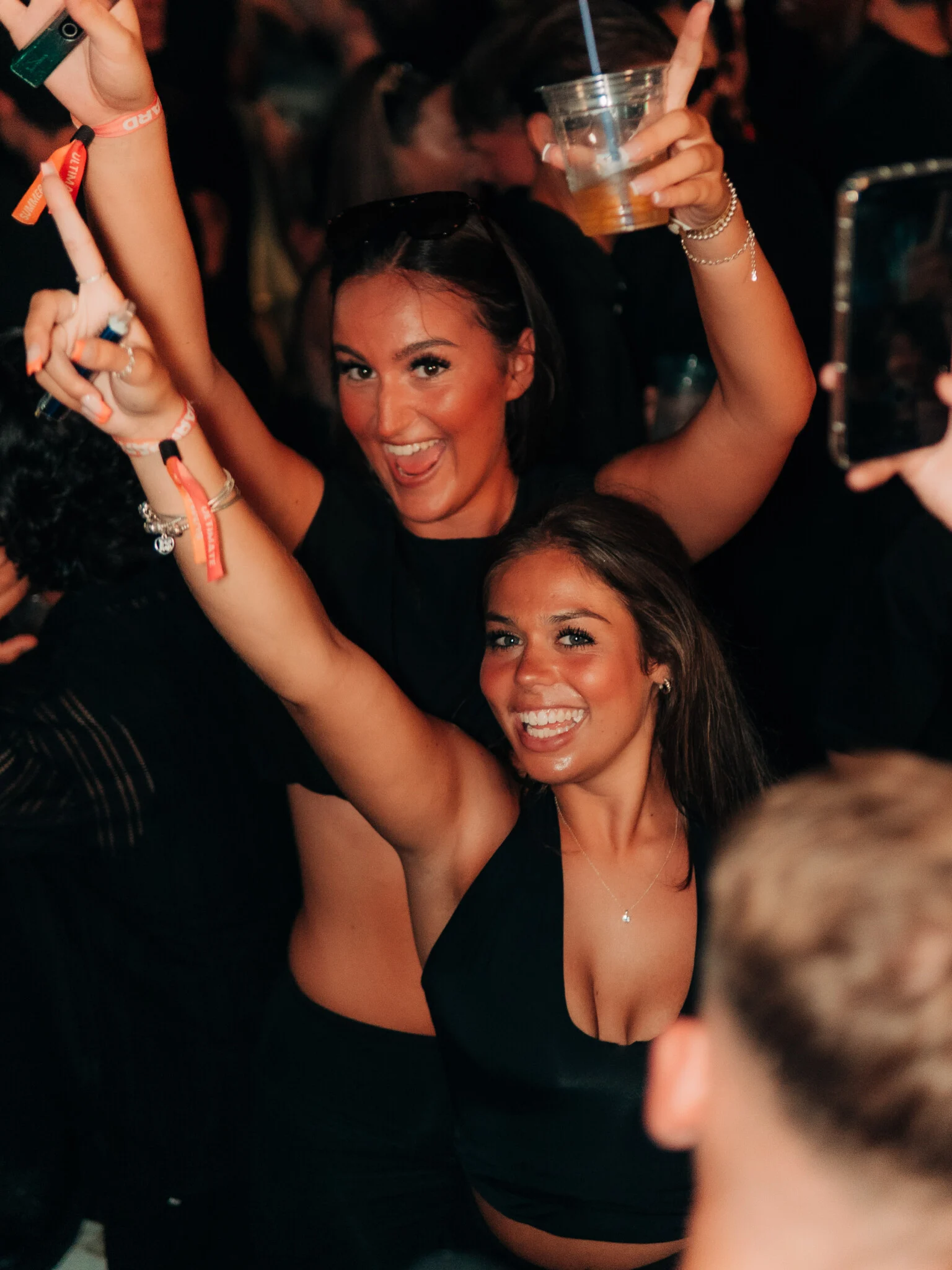 Two girls smiling, dancing, and raising their hands with drinks in the crowd at the Vice Rooftop Party event in Ayia Napa.