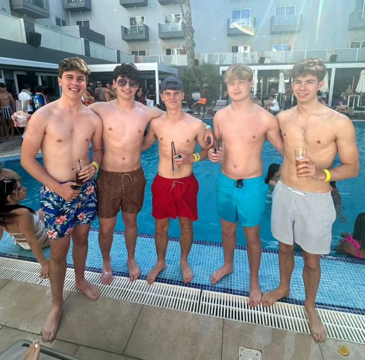 Five lads holding drinks at a busy Ibiza pool party, standing by the pool with summer crowds, sunshine and full daytime party vibes
