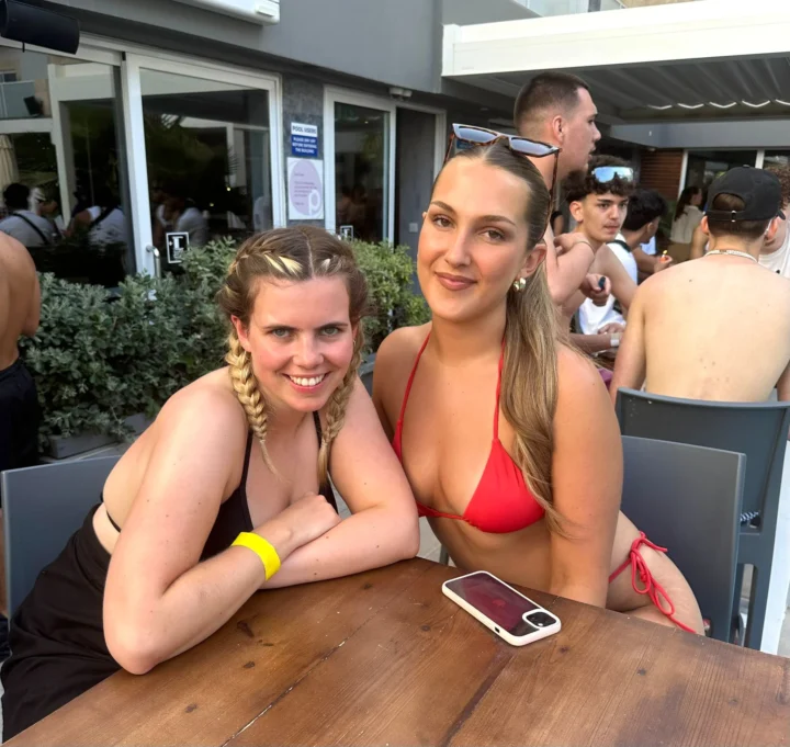 Two girls smiling at a busy Bora Bora Ibiza pool party table, with sunshine, drinks, DJ vibes and a packed daytime crowd