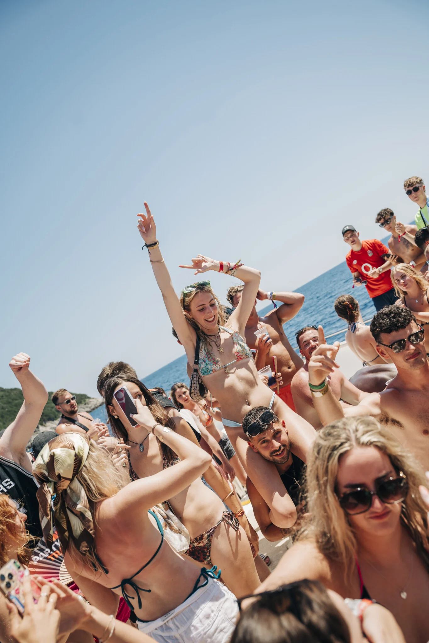 Girl rides on a lad’s shoulders as lads and girls dance on the Kavos Booze Cruise, drinks up and hands in the air