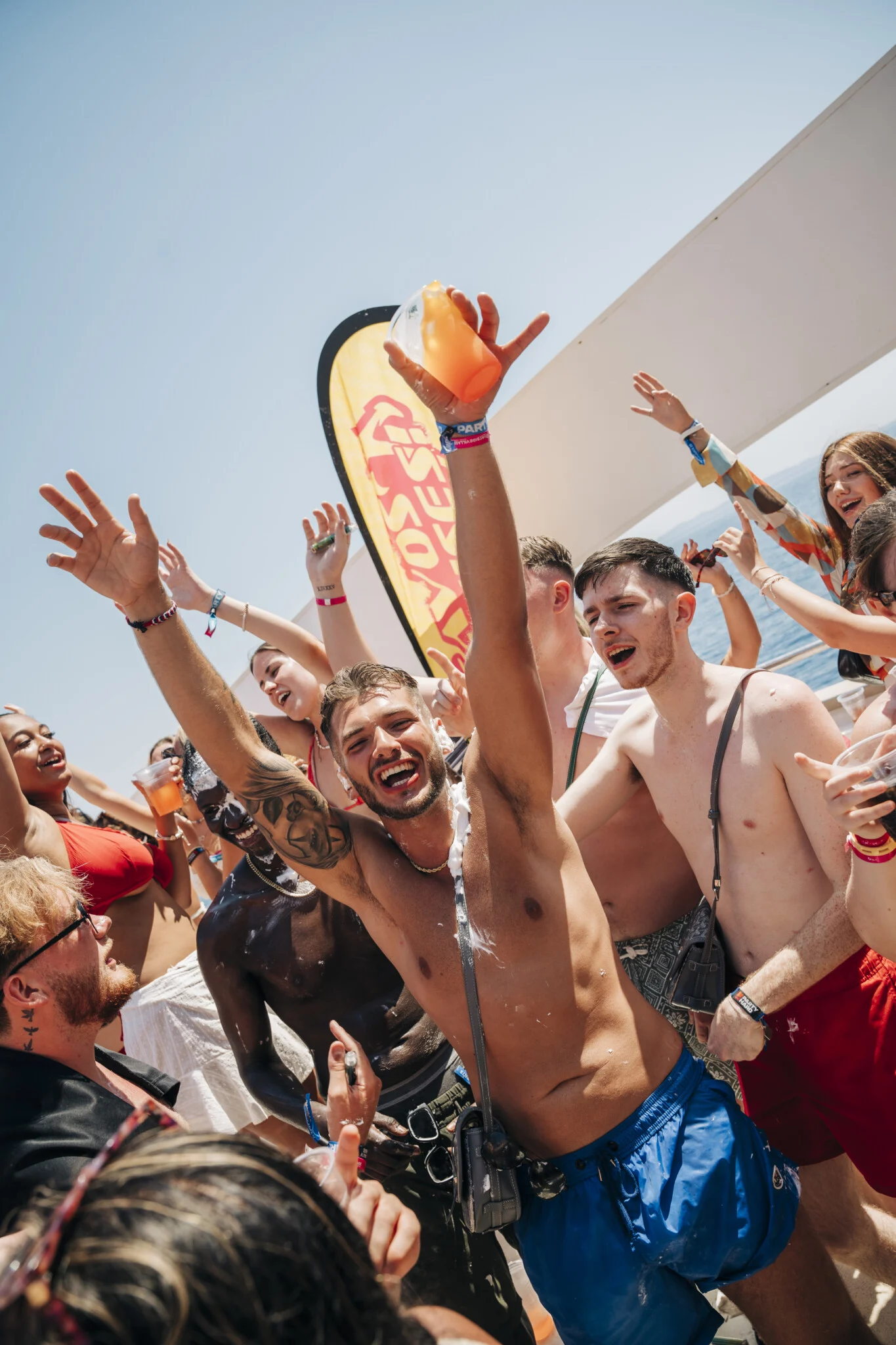 Lads and girls throw drinks in the air on the Kavos Booze Cruise, dancing on deck with hands up and party vibes going off