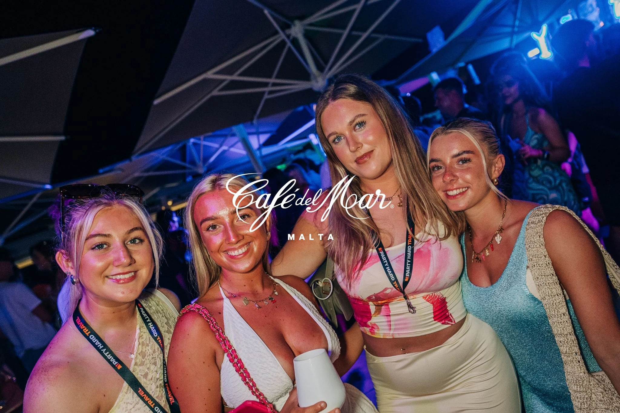 Four girls smiling at Cafe Del Mar Malta sunset party with glowing lights, DJ beats and a buzzing seaside crowd under night skies