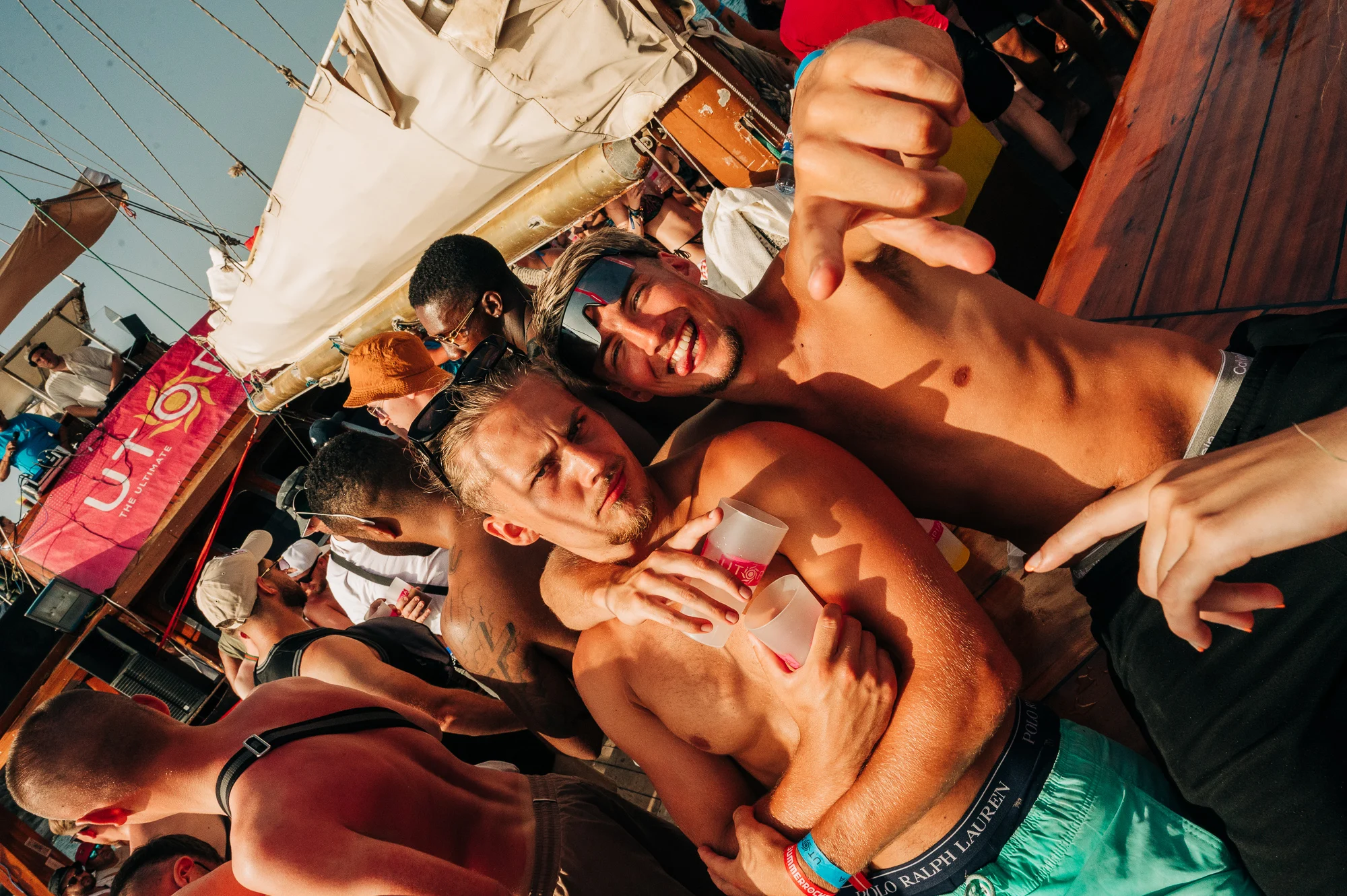 Two lads hyped on a Tenerife boat party, drinks raised, sun blazing and heavy daytime vibes on deck today