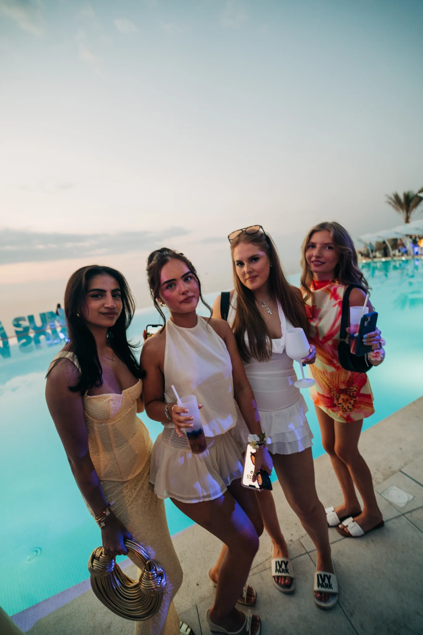 Four girls posing with drinks by the pool at the Toy Room Beach Club Party in Malta during sunset, dressed up for an evening beach club event.