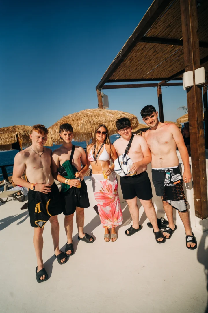 Group of lads and a girl posing with drinks at Toy Room Beach Club in Malta, standing in the sun beside straw parasols and sea views.
