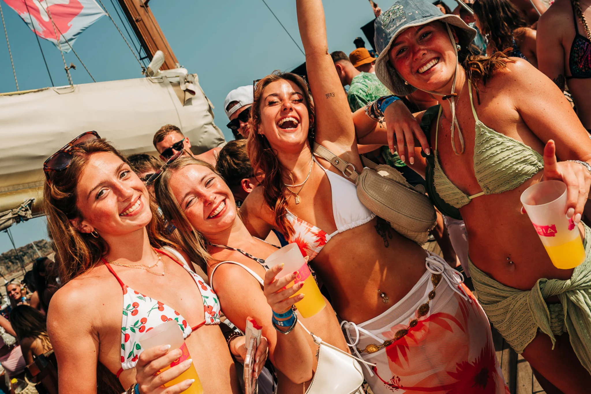 Girls raising drinks on a Tenerife boat party, smiling in swimwear with sunshine, music and packed decks all around