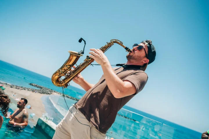 Sax player performing poolside at a Tenerife day party, sunglasses on, ocean backdrop and crowd cooling off