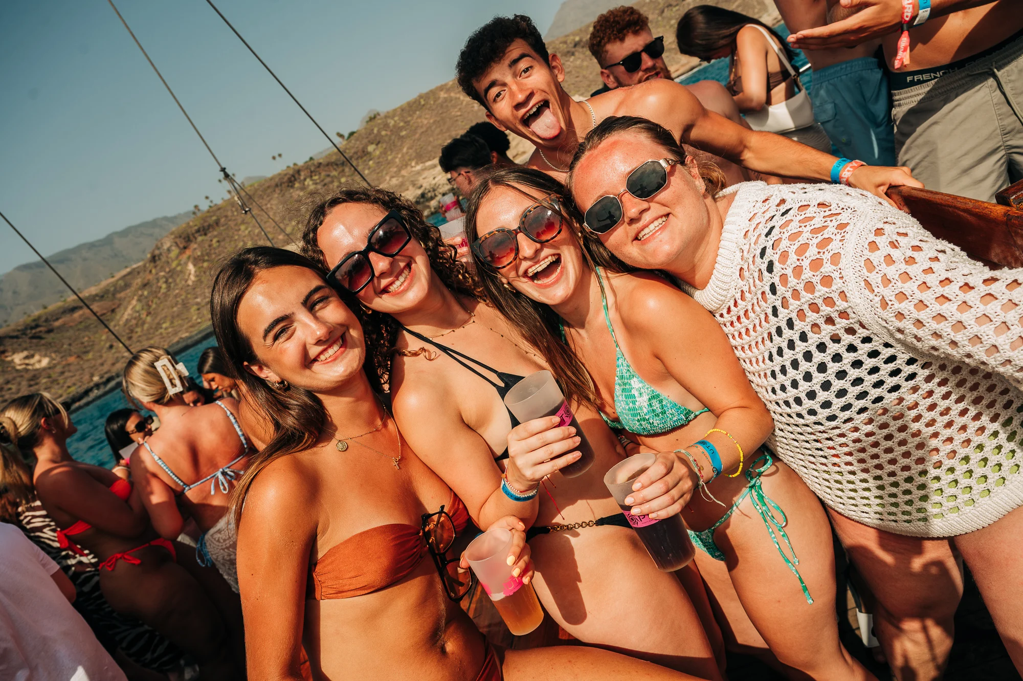 Girls and lads smiling on a Tenerife boat party, drinks in hand, swimwear on deck with sea and hills behind