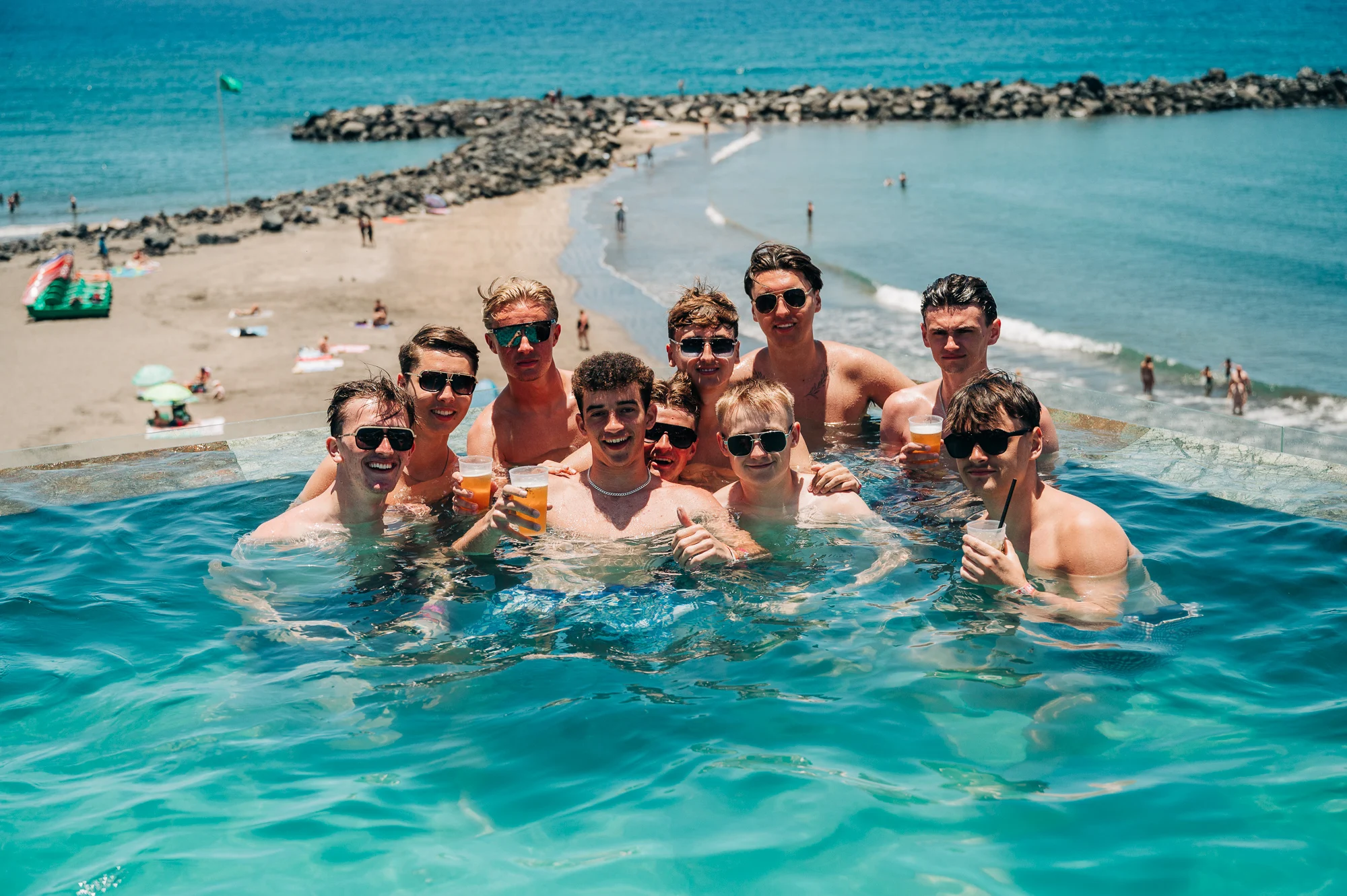 Lads chilling in a pool in Tenerife, beers in hand with ocean views, sunshine and pre-night-out party vibes