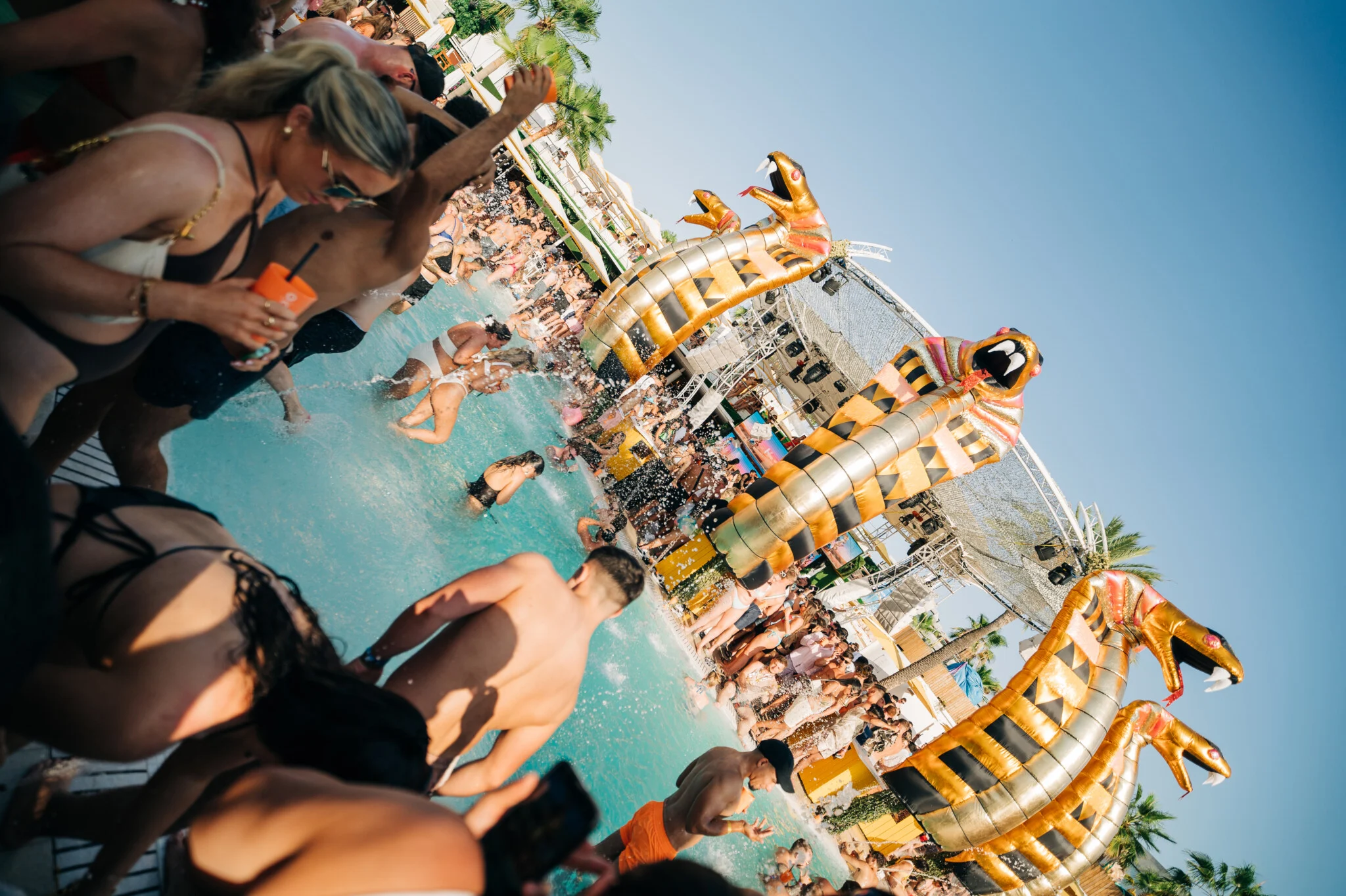 Crowd splashing in the pool beneath giant snake inflatables at O Beach Ibiza during a daytime pool party