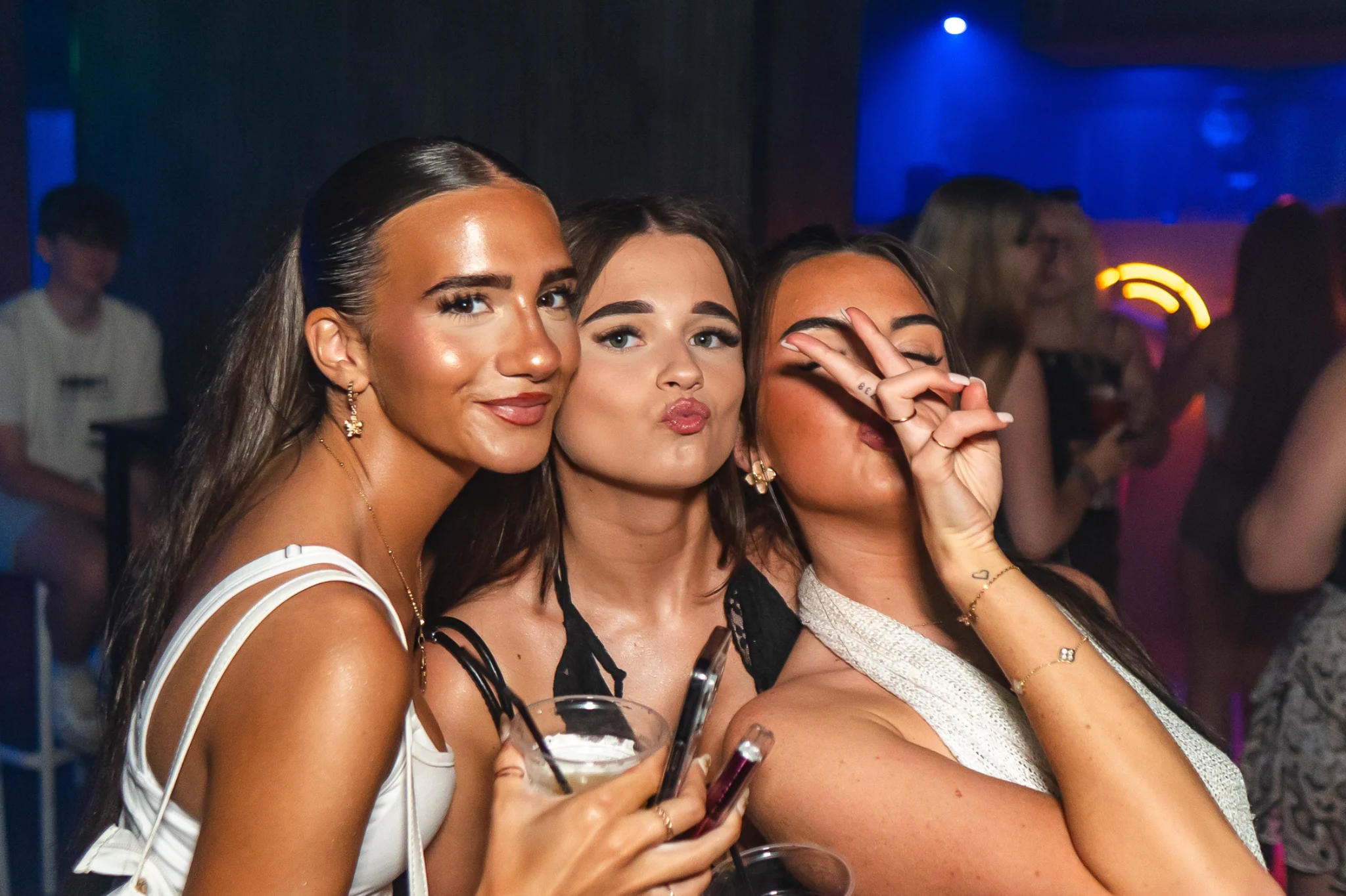 Girls posing on a packed club night, drinks in hand and buzzing with party energy under the neon lights.