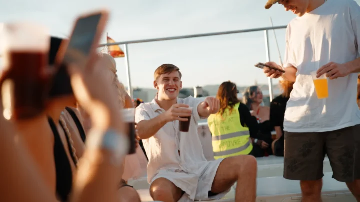 Lads and other guests enjoying drinks on deck at the Barcelona Boat Party in the sunshine, with phones out and the city in the background.