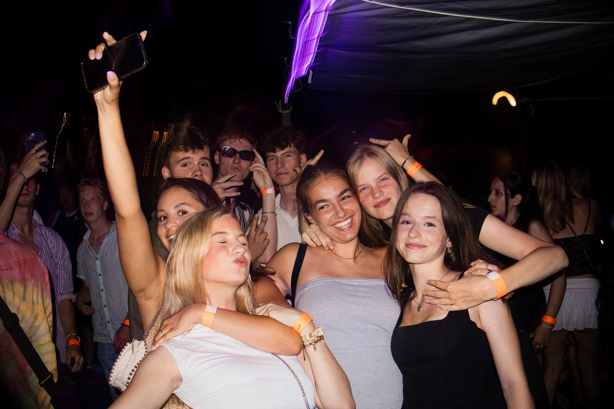 Group of girls and lads posing together at Split After Dark in Croatia, with one girl holding up a phone under purple club lights.