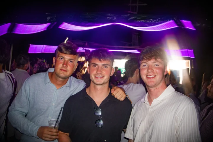 Three lads posing with drinks at Split After Dark in Croatia beneath purple neon lights in a busy club setting.