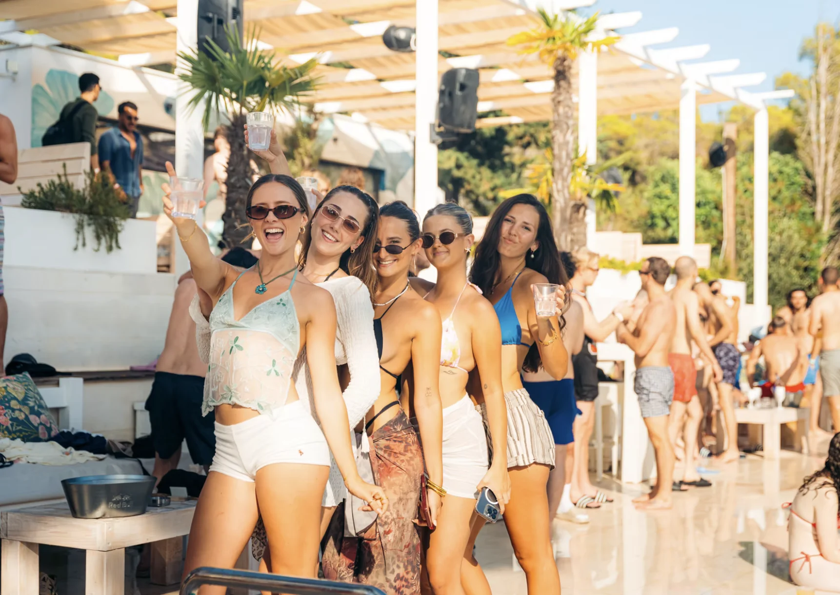 Five girls posing with drinks at the Olife Pool Party in Croatia while guests enjoy the sunshine around the poolside venue