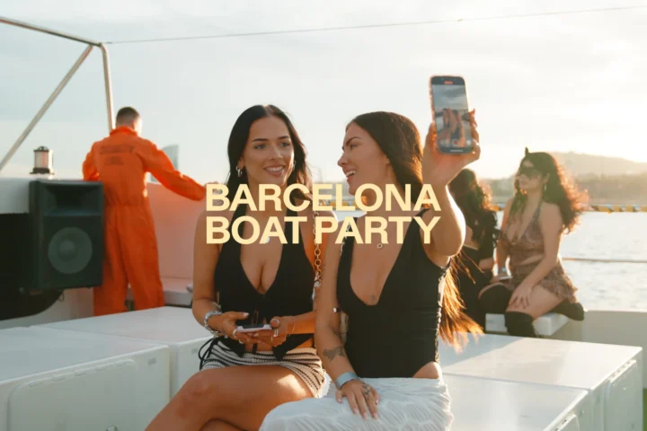 Two girls sitting on board at the Barcelona Boat Party, taking a phone photo in the sun with the event logo across the image.