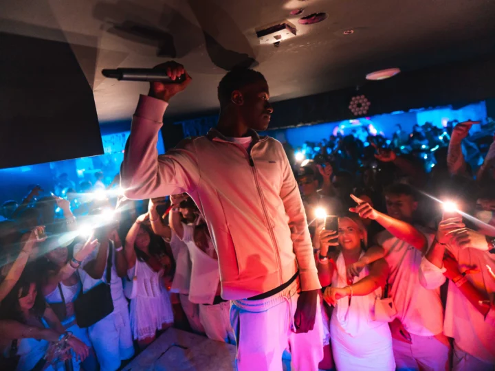 MC holding a microphone on stage as the crowd in white outfits film and cheer at Napa Xtra in Ayia Napa