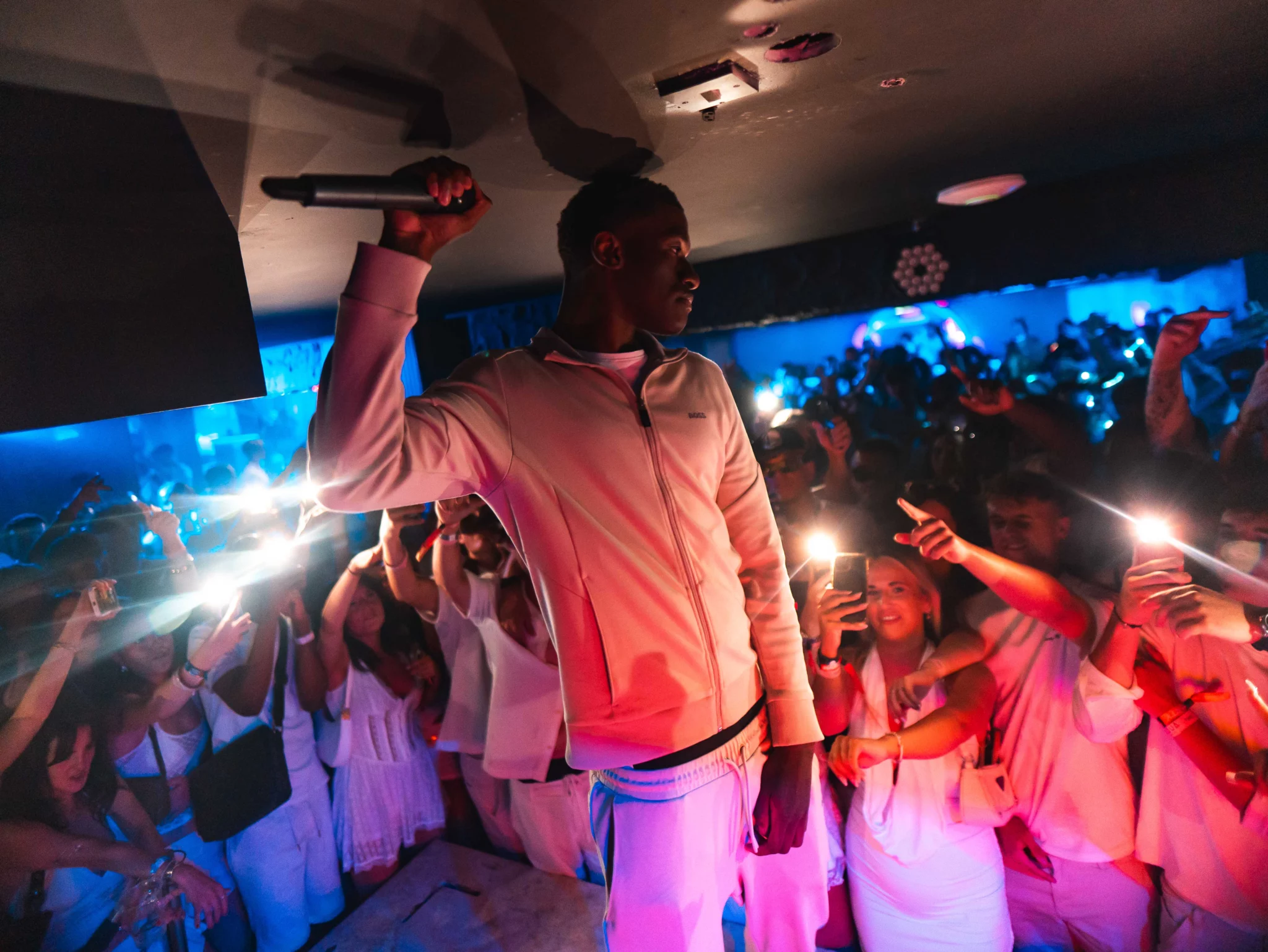 MC holding a microphone on stage as the crowd in white outfits film and cheer at Napa Xtra in Ayia Napa